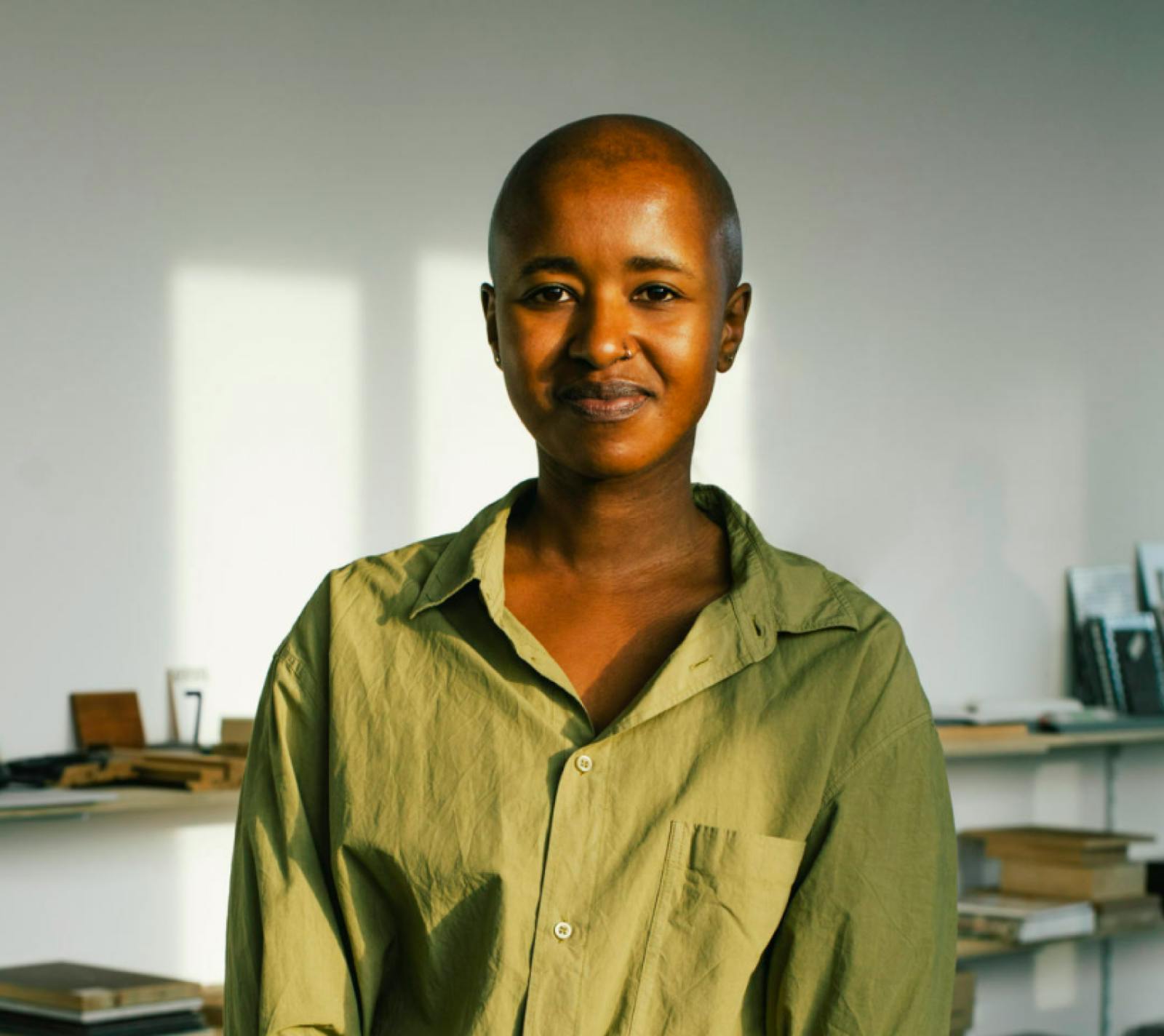 Portrait of a black woman with saved head and a nose piercing looking to the camera. She wears a green shirt and she's in front of a white wall with some shelves blurred in the background